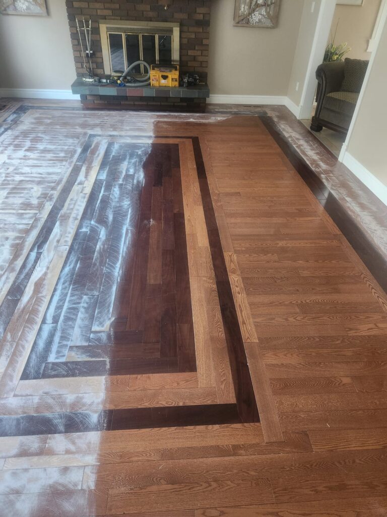 Worn original finish on a living room floor, sanding dust collecting near the brick fireplace